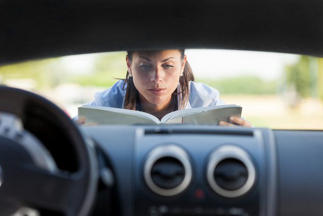 caucasian woman reads manual while under hood of car